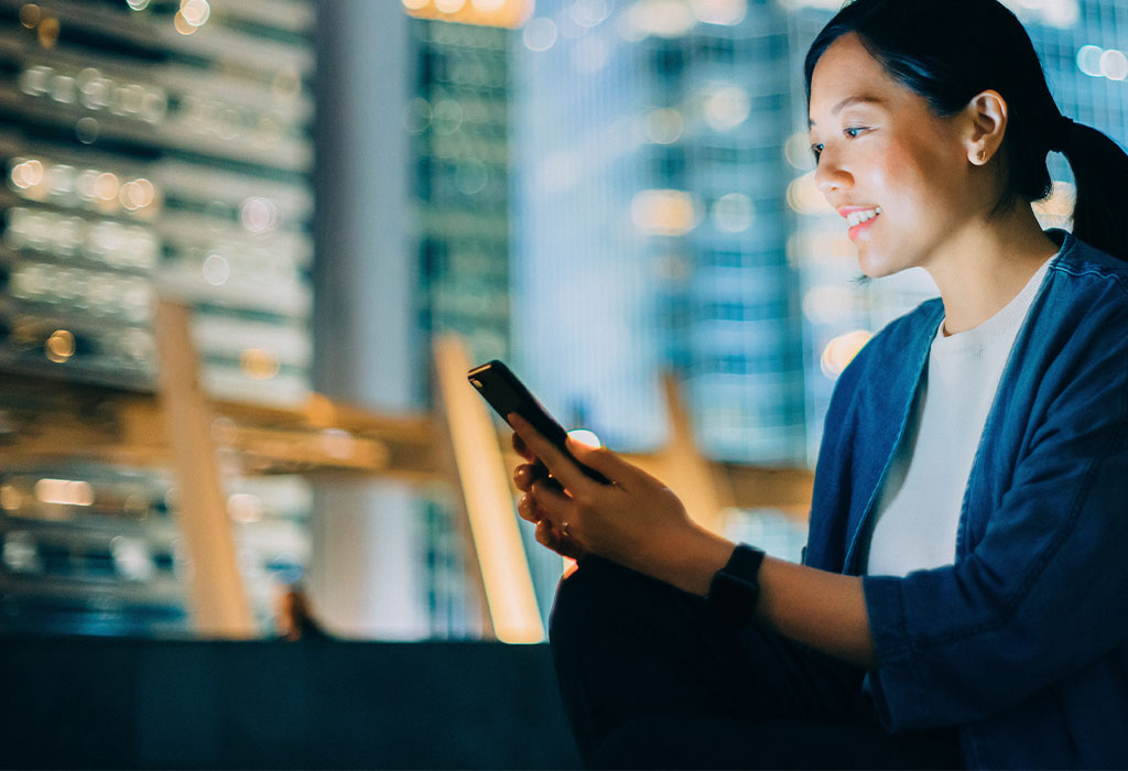 A young woman sitting outside at night looking at a mobile phone with city lights in the background